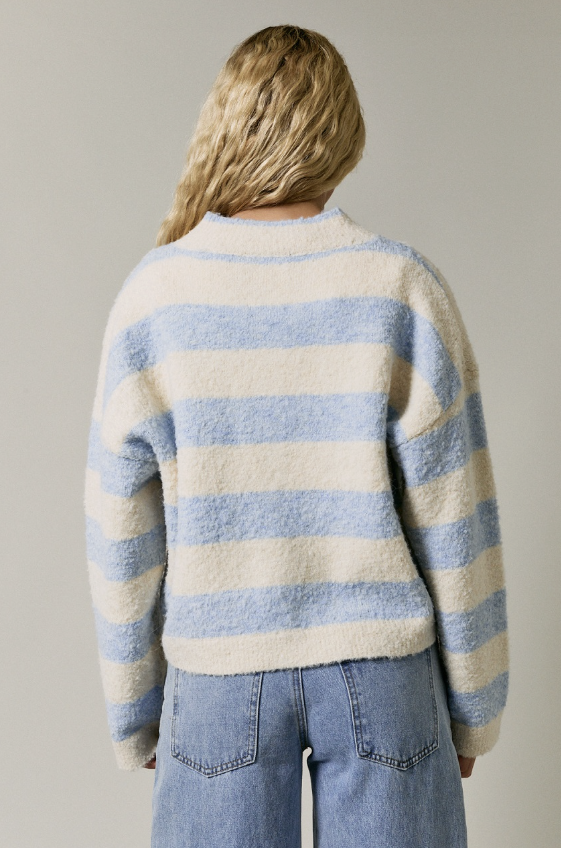 A woman viewed from behind wearing a fuzzy knit sweater with horizontal light blue and cream stripes, paired with light-wash blue jeans, against a plain light gray studio background.