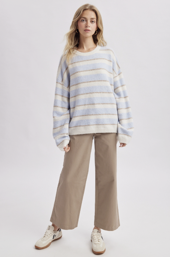A woman modeling a fuzzy, oversized long-sleeve sweater with horizontal stripes in light blue, beige, and white, paired with wide-leg beige trousers and white sneakers, against a plain light gray studio background.