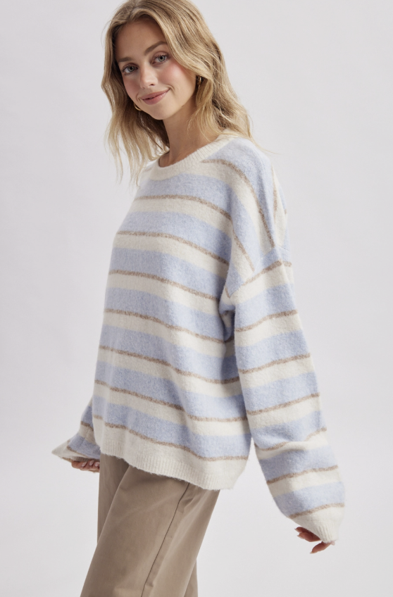 A woman wearing a fuzzy knit crewneck sweater with horizontal stripes in light blue, cream, and beige, paired with beige trousers, against a light gray studio background.