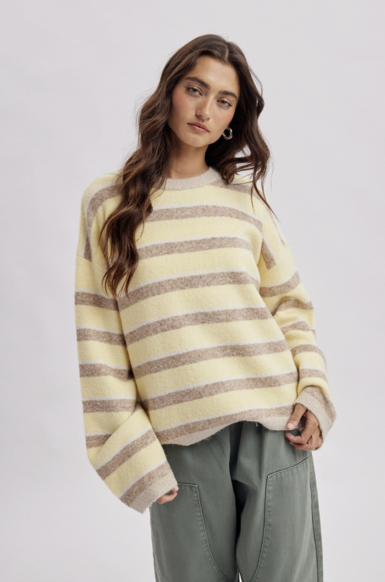 A woman wearing a cream and taupe striped knit sweater with voluminous long sleeves, paired with olive green pants, against a light gray studio background.