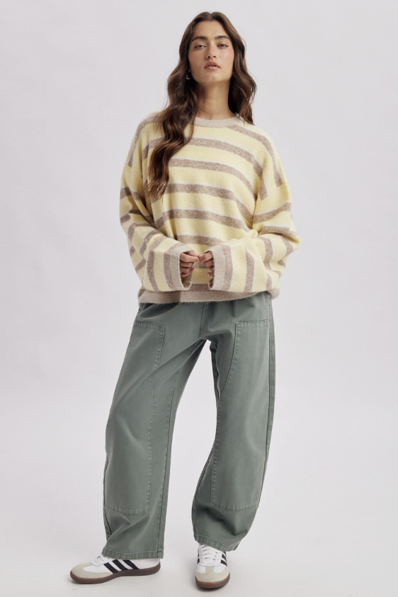 A woman modeling a cream and taupe striped fuzzy knit sweater, paired with wide-leg sage green pants and white sneakers, against a light gray studio background.