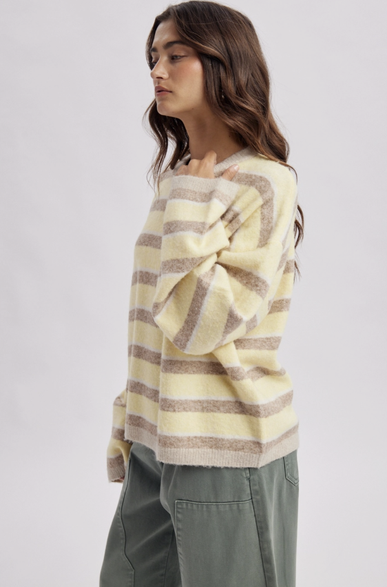 A woman wearing a cream and taupe striped fuzzy knit sweater with wide sleeves, paired with olive green pants, shown from a side profile against a light gray studio background.