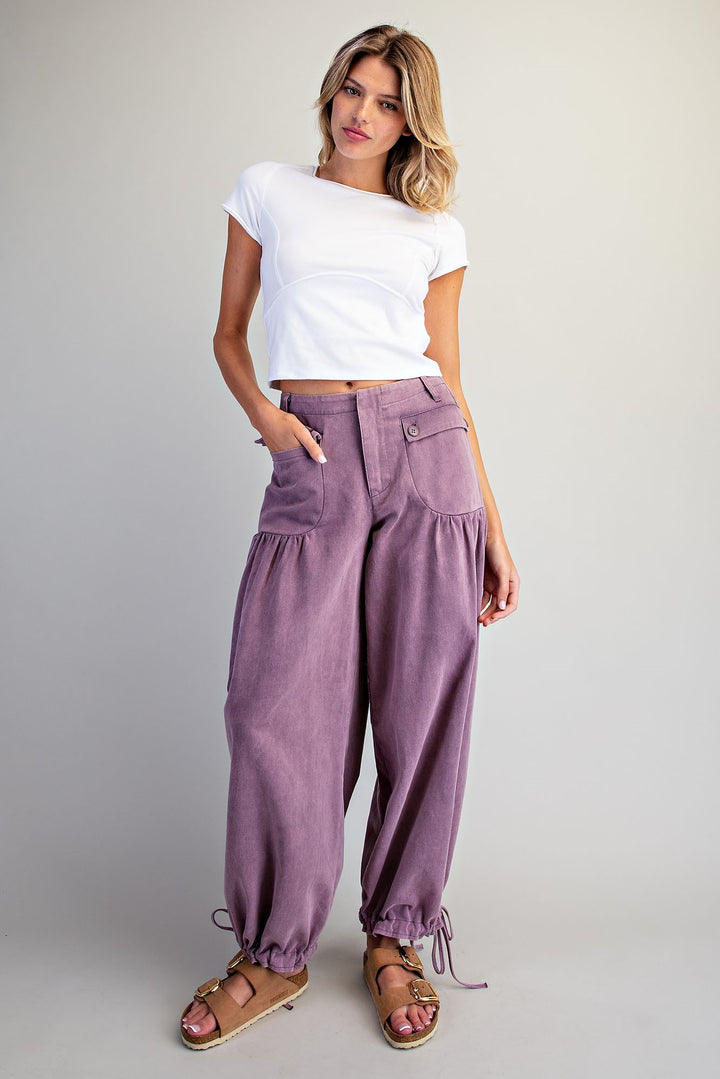 A woman modeling a pair of loose, high-waisted mauve-purple corduroy pants with drawstring ankle cuffs, paired with a white t-shirt and brown sandals, against a light gray studio background.