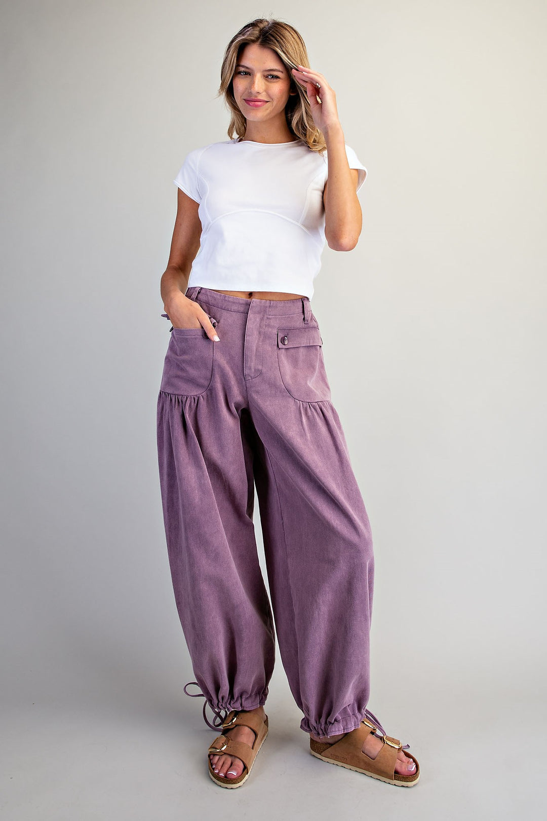 A woman modeling loose, wide-leg purple pants with a white short-sleeve crop top and brown sandals, standing against a plain light gray studio background.