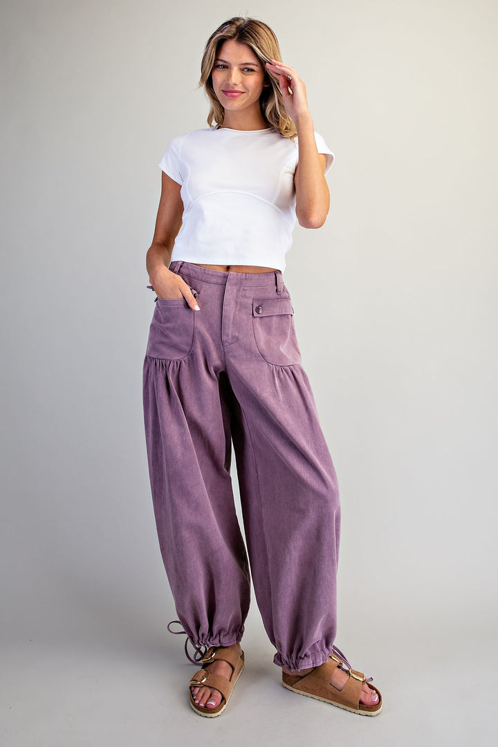A woman modeling loose, wide-leg purple pants with a white short-sleeve crop top and brown sandals, standing against a plain light gray studio background.