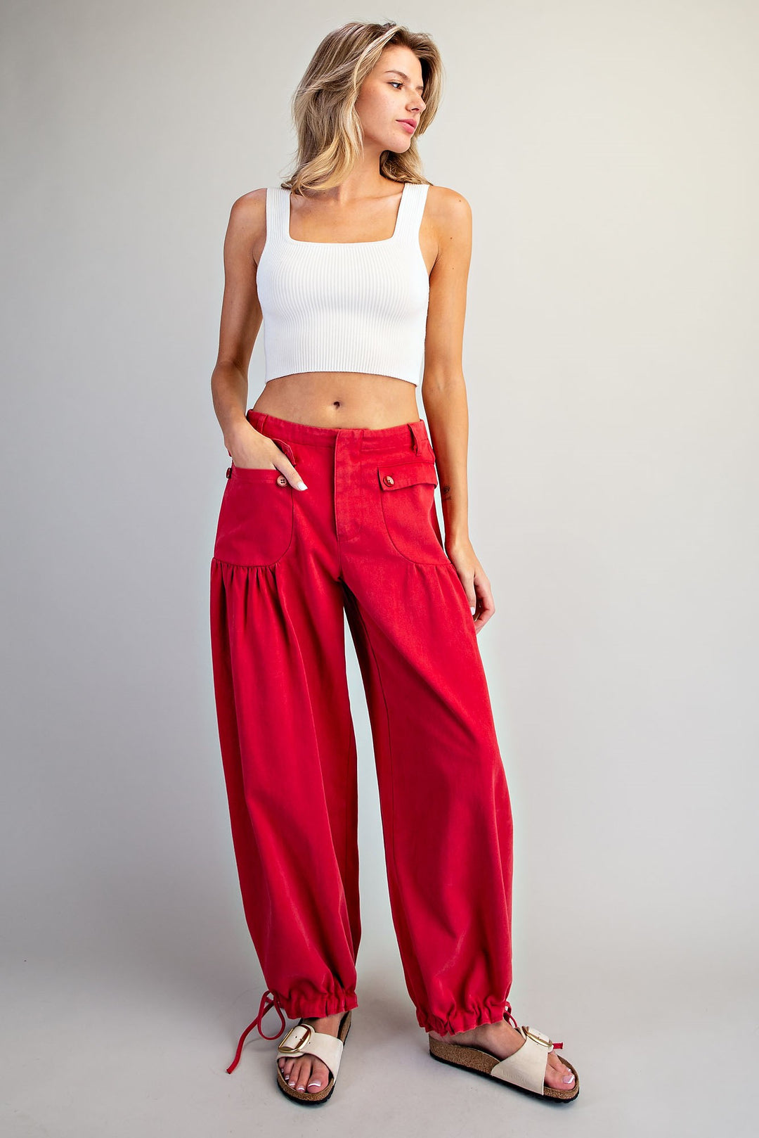 A woman modeling bright red wide-leg cropped pants with gathered harem-style cuffs, paired with a white crop top and beige sandals, against a light gray studio background.