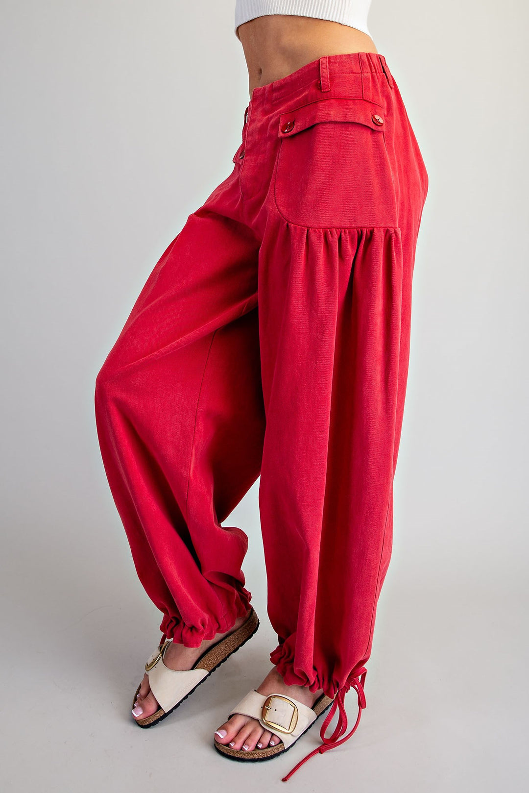 A person wearing bright red wide-leg drawstring pants with gathered side details and white slide sandals, shown from the waist down against a light gray studio background.