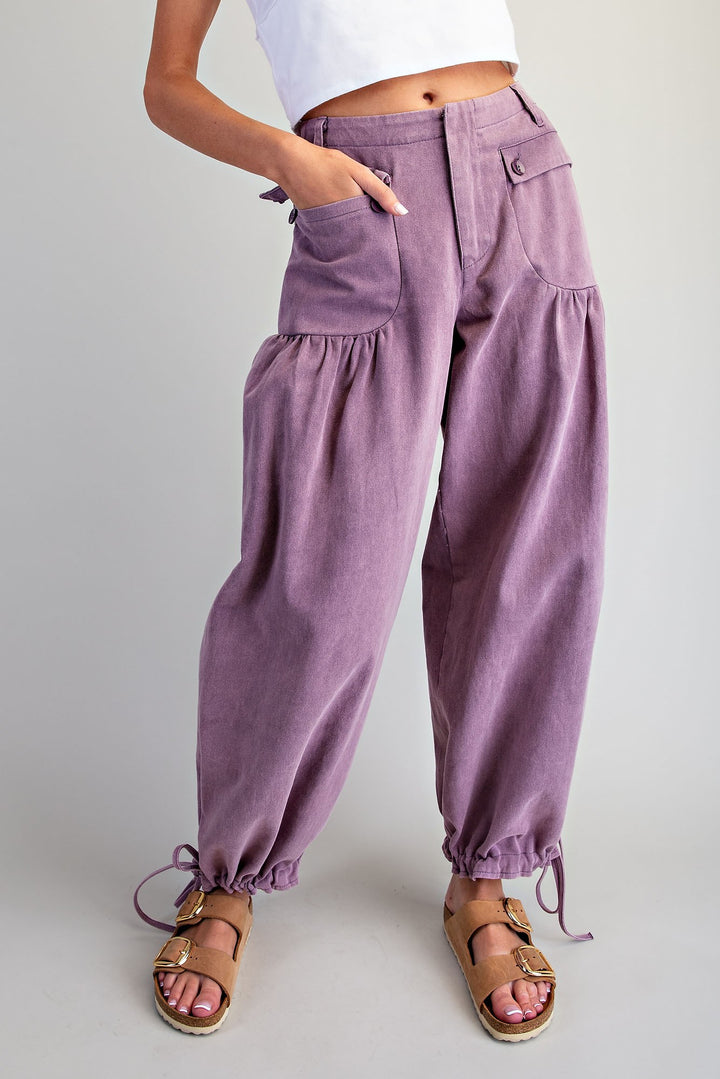 A person wearing loose, balloon-style lavender pants with gathered side pockets and drawstring ankle cuffs, paired with tan sandals and a white cropped top against a light gray studio background.