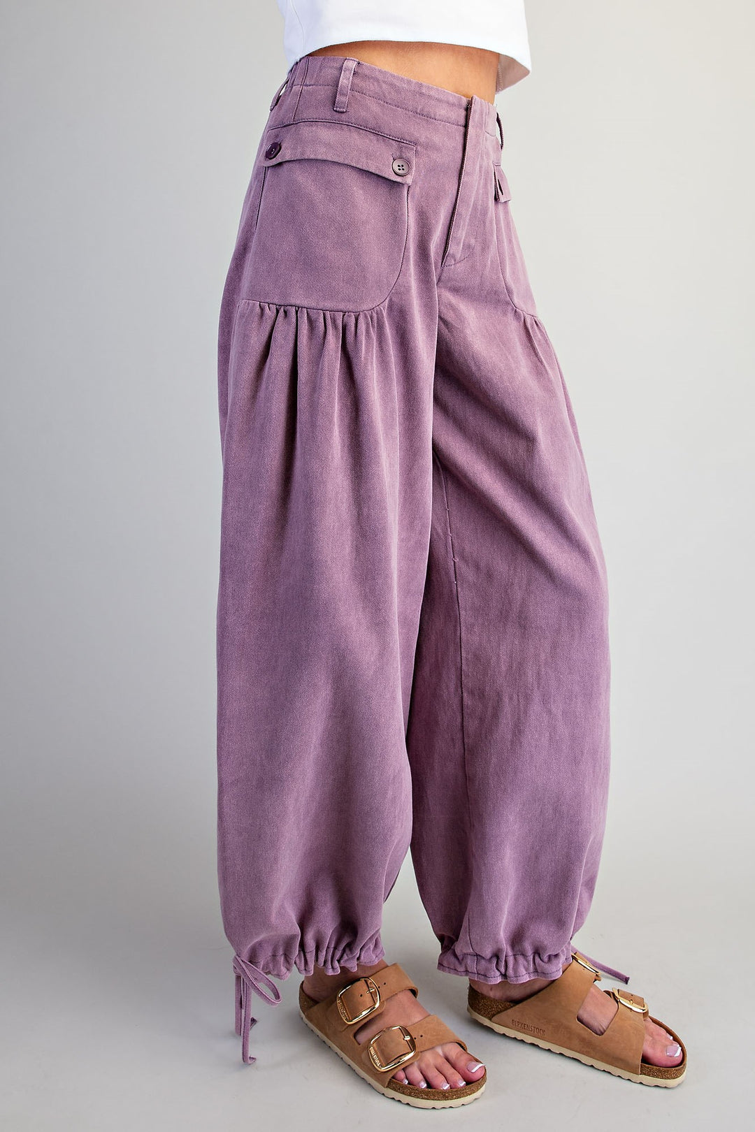 A person wearing loose, balloon-style lavender corduroy pants with gathered side pockets and drawstring hems, paired with tan double-strap sandals against a plain light gray studio background.