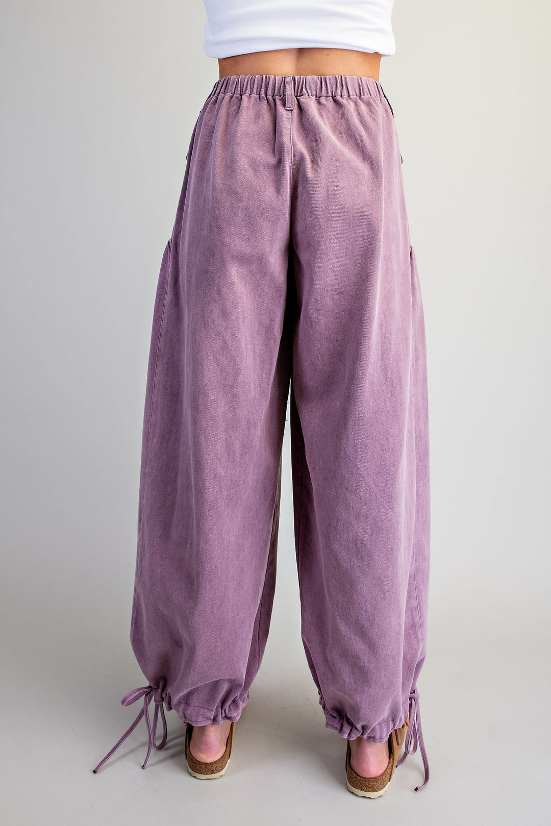 A person shown from the waist down wearing loose, wide-leg mauve-purple corduroy pants with an elastic waistband and drawstring ankle cuffs, paired with brown wedge sandals against a plain light gray studio background.