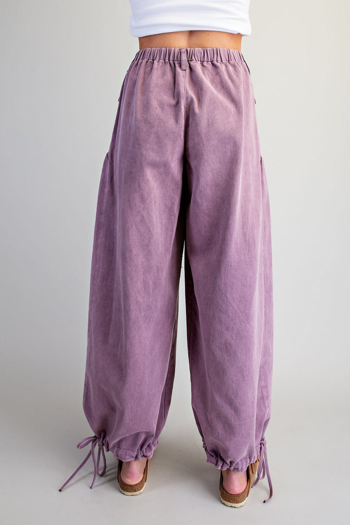 A person shown from the waist down wearing loose, wide-leg mauve-purple corduroy pants with an elastic waistband and drawstring ankle cuffs, paired with brown wedge sandals against a plain light gray studio background.