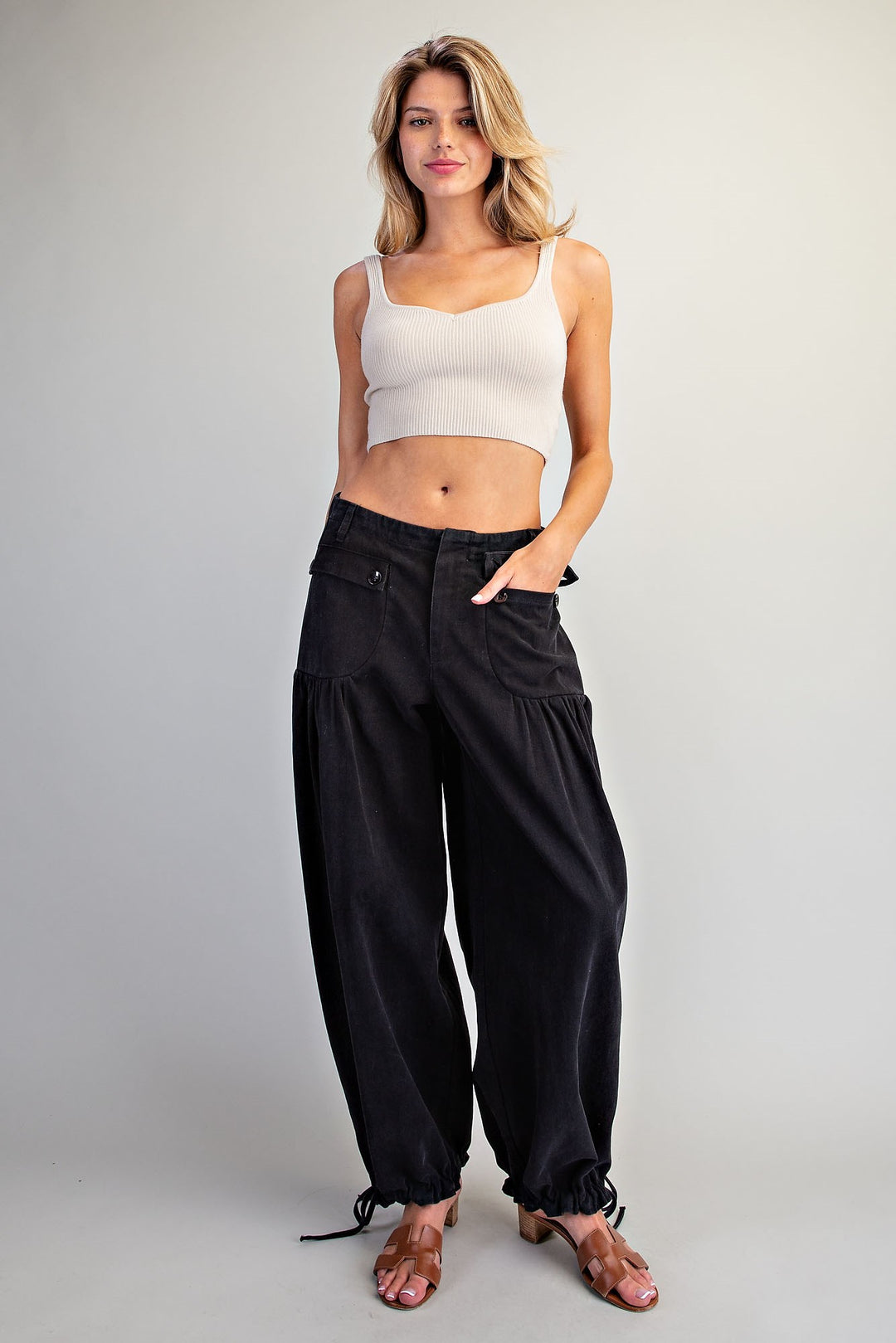 A woman modeling black wide-leg harem pants with drawstring ankle cuffs, paired with a white ribbed crop top and brown heeled sandals, against a light gray studio background.