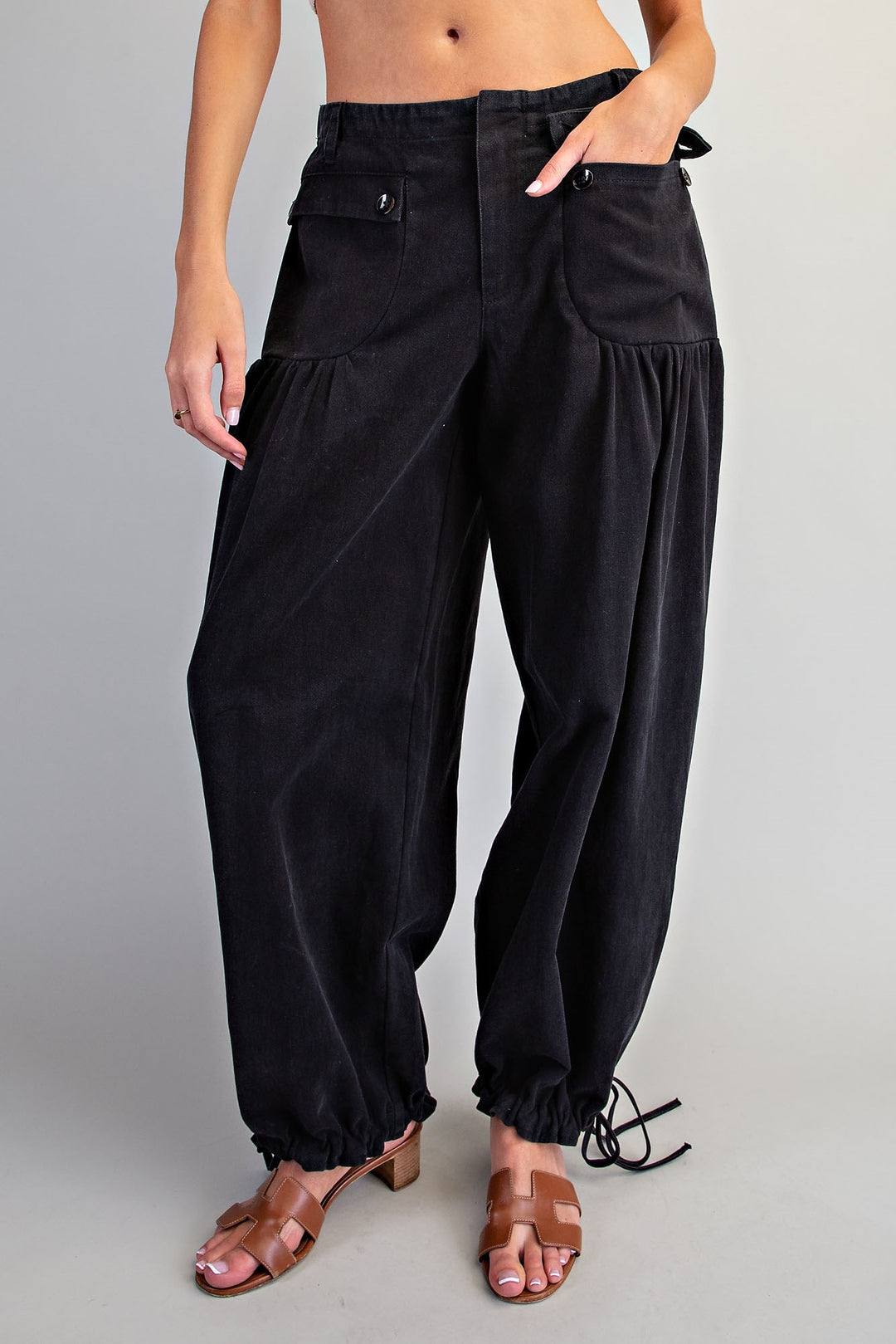 Black wide-leg corduroy pants with gathered side pockets and elastic ankle cuffs, shown on a model wearing brown sandals against a light gray studio background.