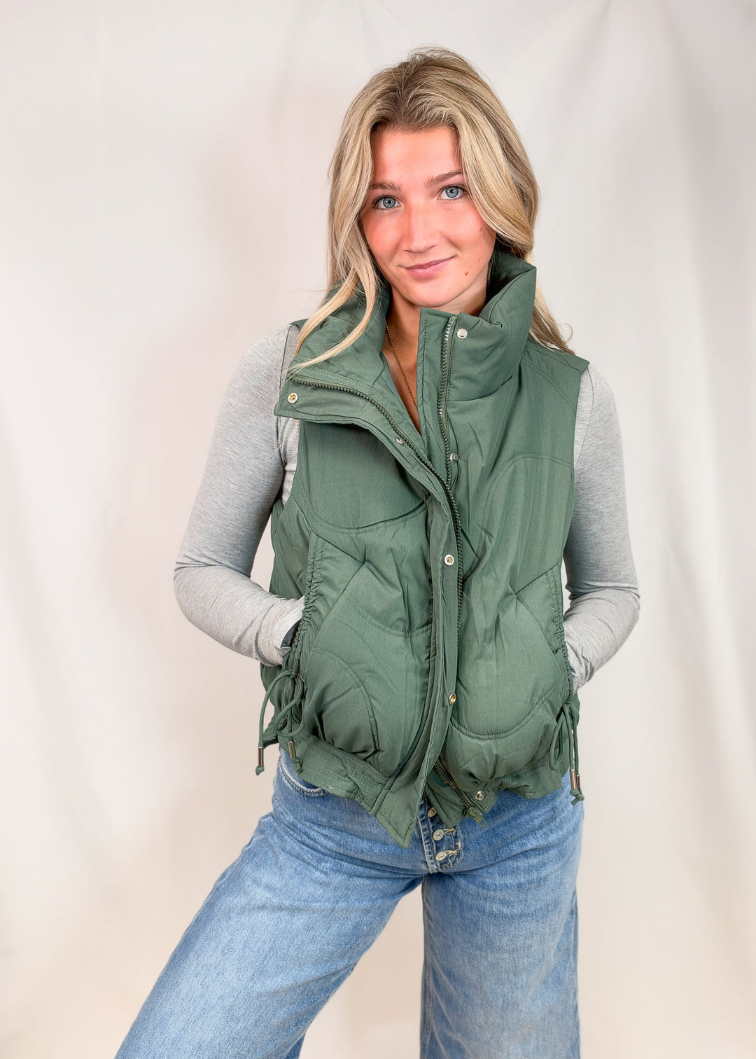 A woman wearing an olive green puffer vest over a gray long-sleeve shirt and blue jeans, standing against a light beige studio backdrop.