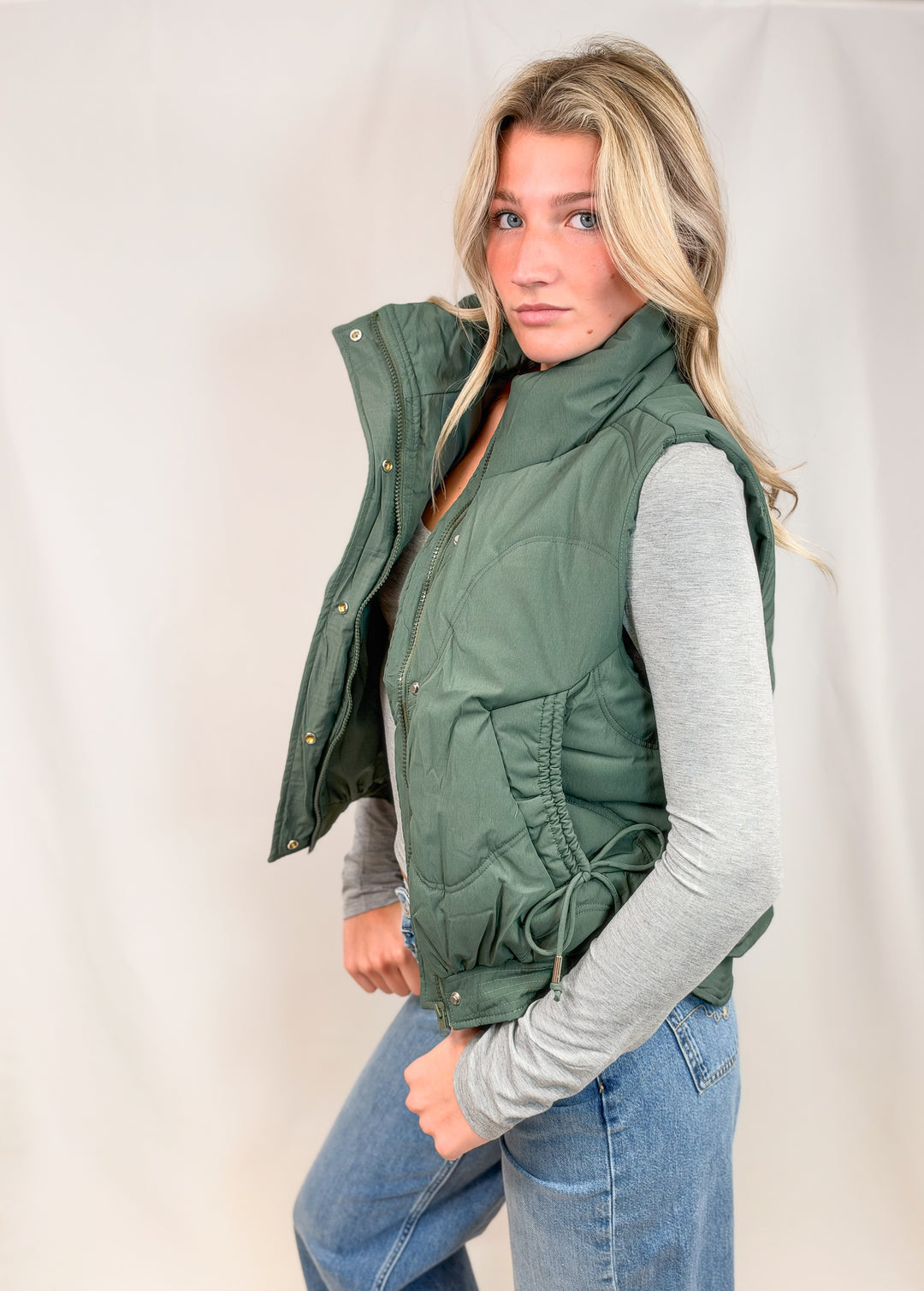 A woman wearing an olive green sleeveless puffer vest over a light gray long-sleeve shirt and blue jeans, standing against a light beige studio backdrop.
