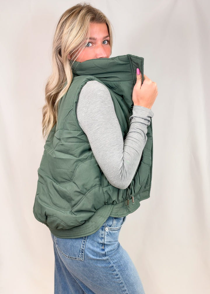 A woman wearing an olive green puffer vest over a light gray long-sleeve shirt and blue jeans, holding the high collar of the vest up with one hand against a plain light background.