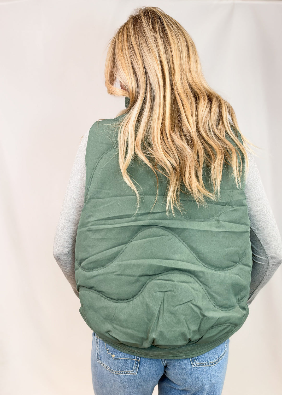 A woman viewed from behind wearing a sage green quilted puffer vest over a light gray long-sleeve shirt and blue jeans, set against a plain light gray studio backdrop.
