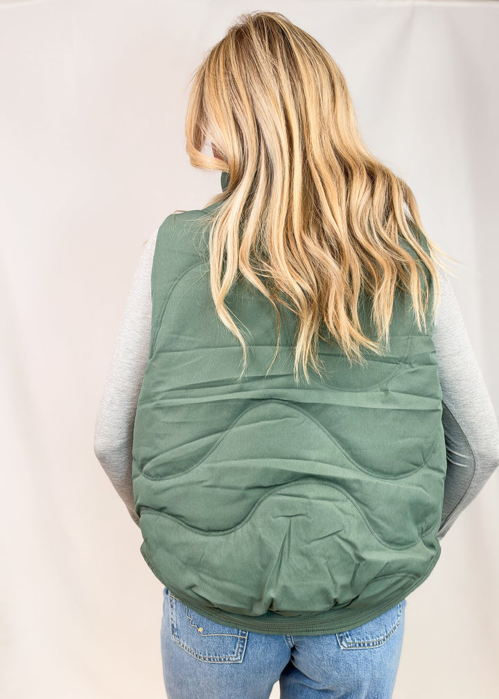A woman viewed from behind wearing a sage green quilted puffer vest over a light gray long-sleeve shirt and blue jeans, set against a plain light gray studio backdrop.