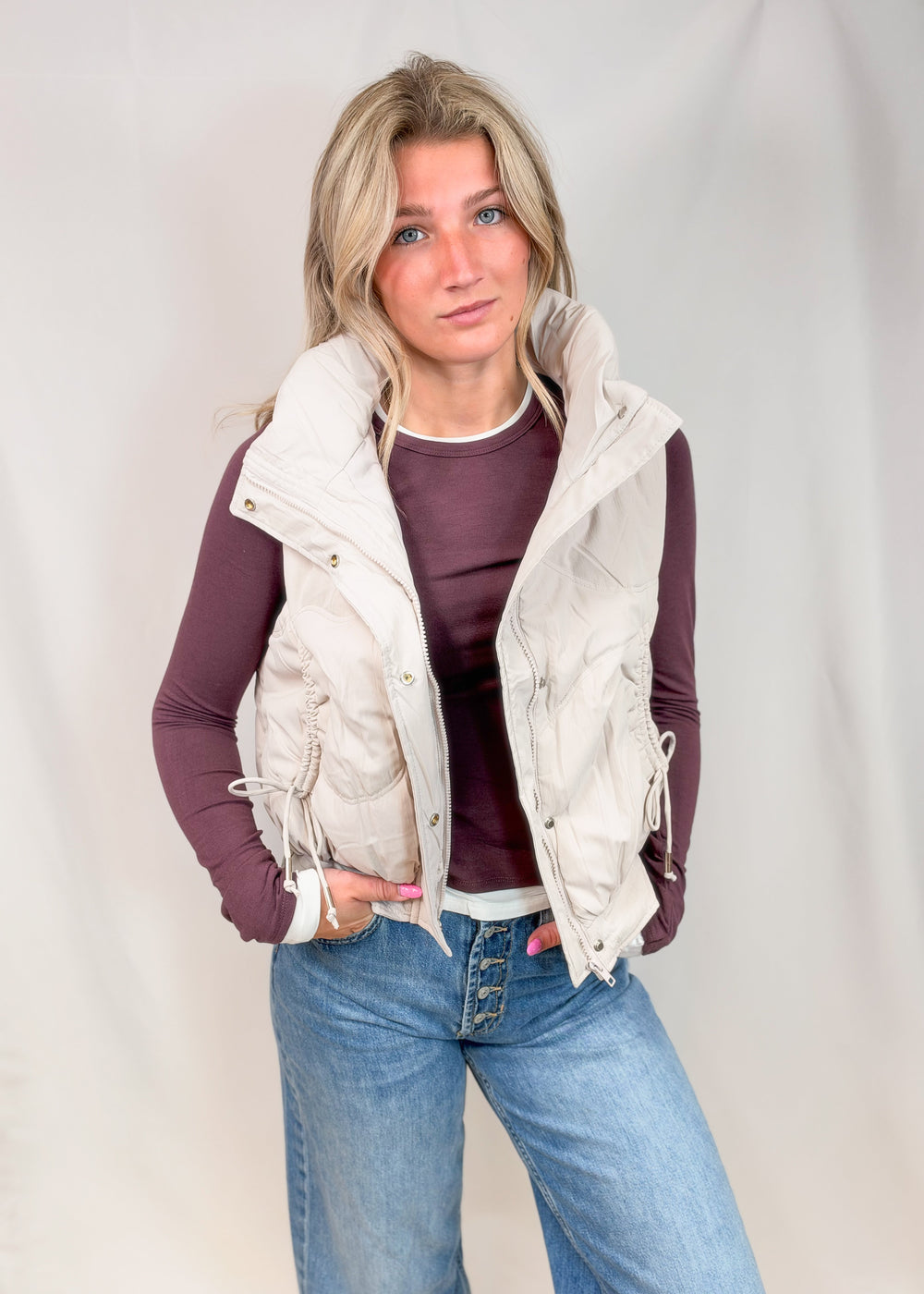 A woman modeling a sleeveless cream quilted puffer vest over a dark plum long-sleeve top and blue jeans, standing against a light gray studio backdrop.