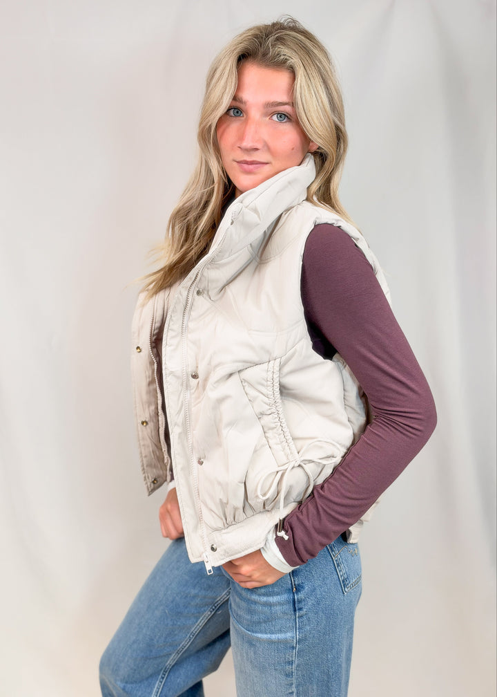 A woman modeling a light beige sleeveless quilted vest over a dark plum long-sleeve shirt and blue jeans, standing against a light gray fabric backdrop.