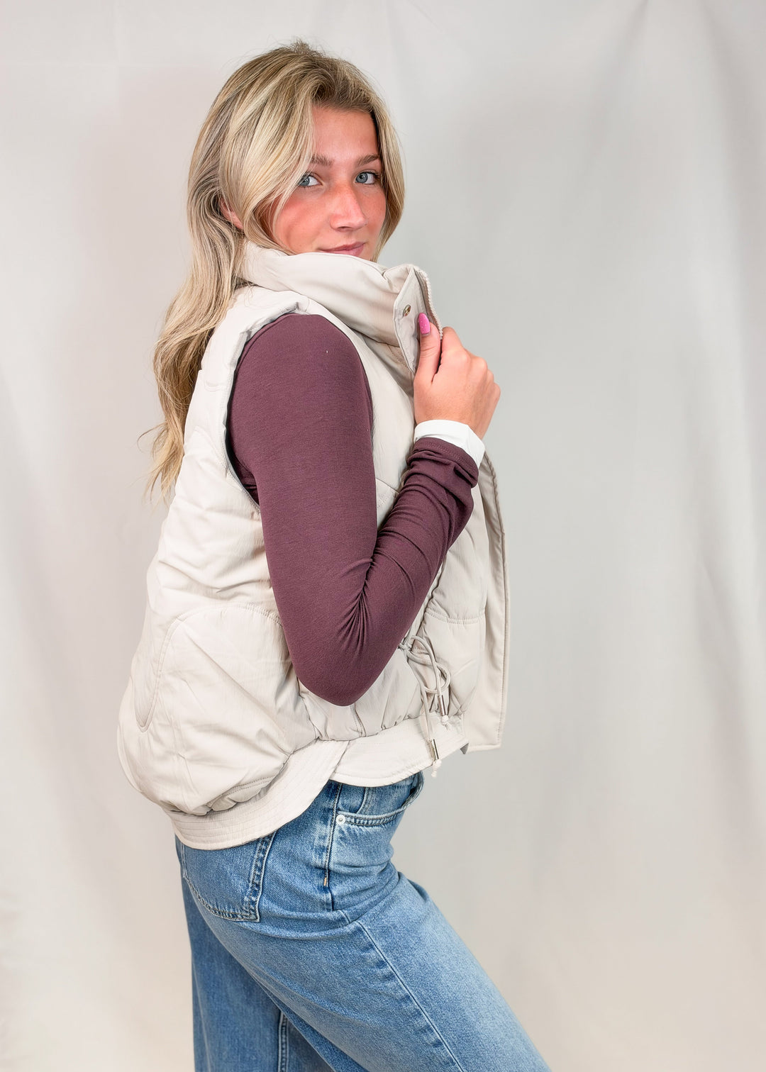 A woman modeling a light beige sleeveless puffer vest over a dark plum long-sleeve top and blue jeans, posed against a light gray fabric backdrop.