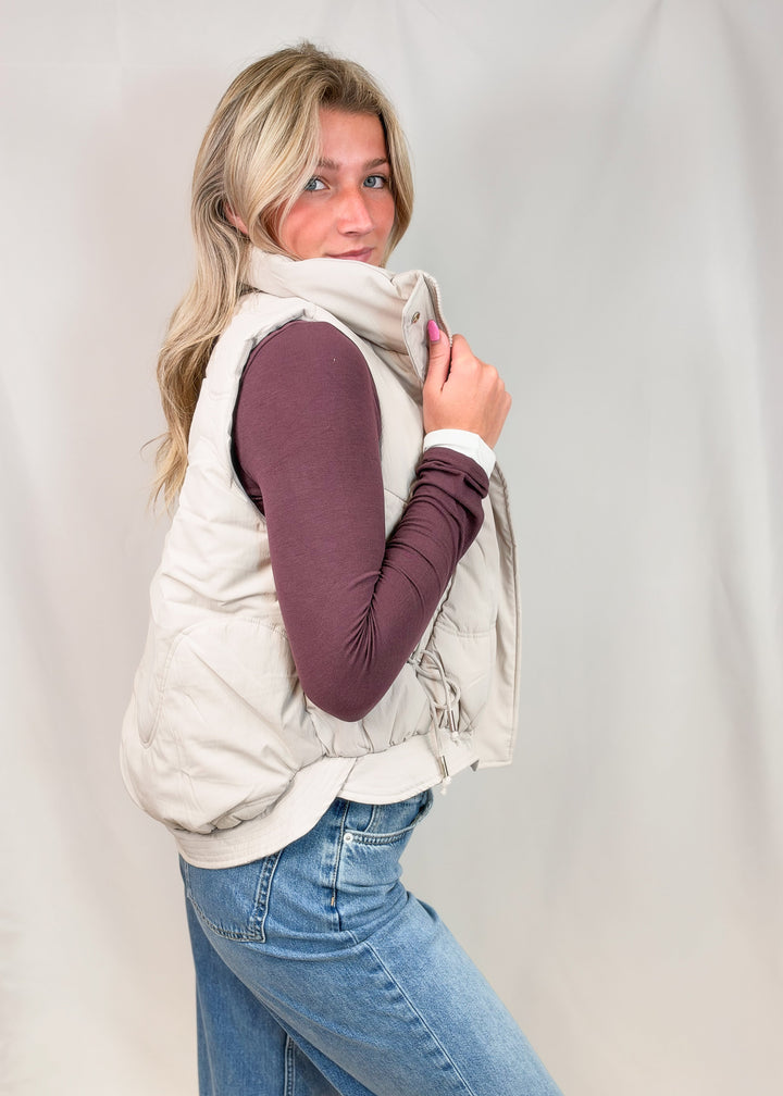 A woman modeling a light beige sleeveless puffer vest over a dark plum long-sleeve top and blue jeans, posed against a light gray fabric backdrop.