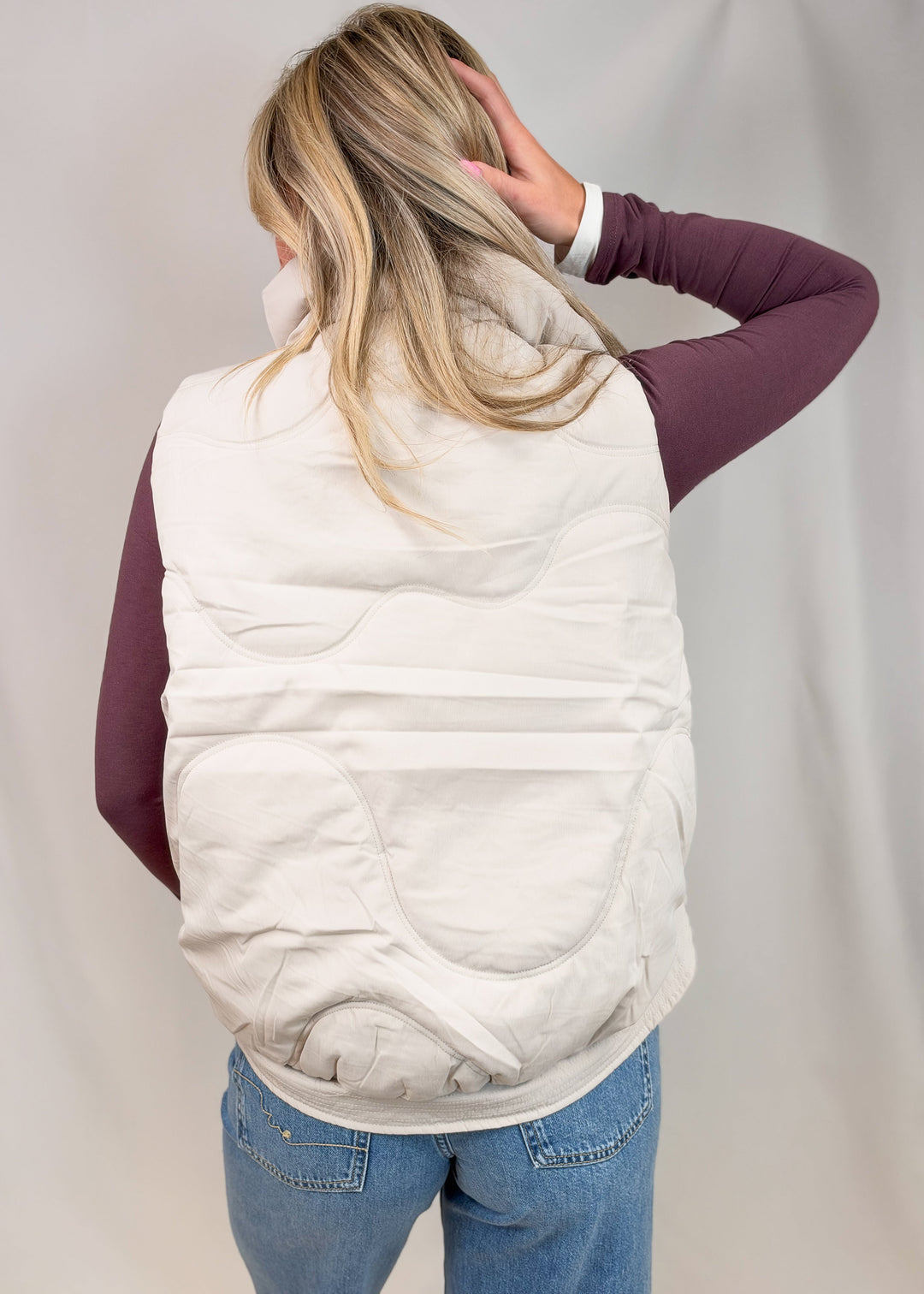 A woman viewed from behind wearing a cream quilted puffer vest over a dark plum long-sleeve shirt and blue jeans, standing against a light gray fabric backdrop.