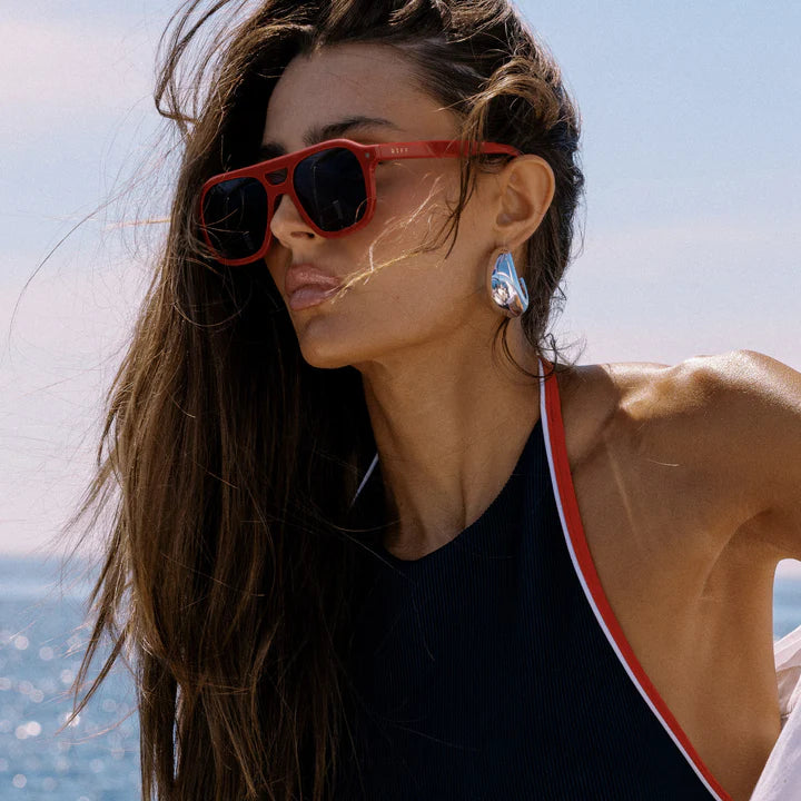 Woman wearing red sunglasses and a black swimsuit with a red stripe, standing against a blue sky.