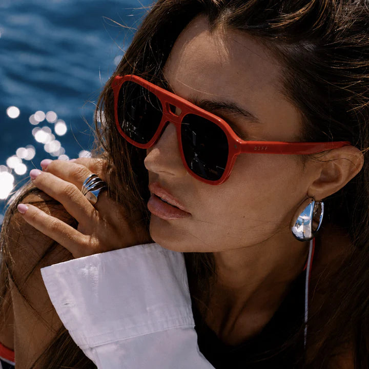 Woman wearing red sunglasses with a blurred background of water and sunlight.