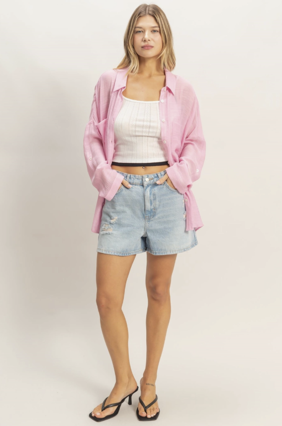 Woman wearing a pink shirt, white top, and denim shorts on a plain background
