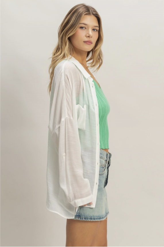 Woman wearing a white sheer cardigan over a green top and denim shorts on a plain background