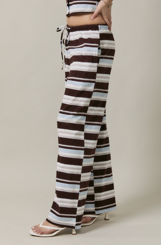 A person wearing wide-leg drawstring pants with horizontal brown, white, and light blue stripes, paired with a matching striped crop top and white heeled sandals, shown against a plain beige studio background.