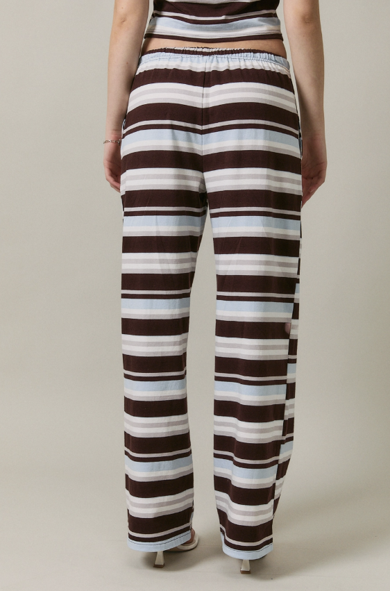A person shown from the back wearing loose, wide-leg striped pants with horizontal bands in dark brown, white, and light blue on a plain beige studio background.