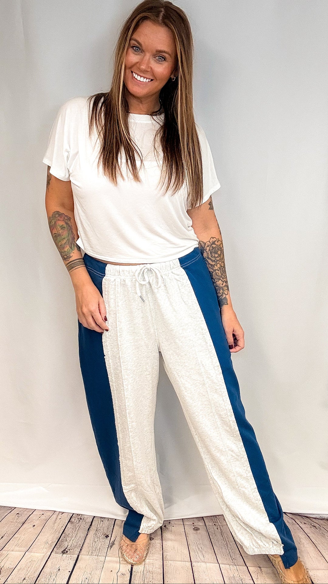 A woman modeling a white t-shirt and white wide-leg sweatpants with blue side panels, standing against a white studio backdrop with a light wood floor.