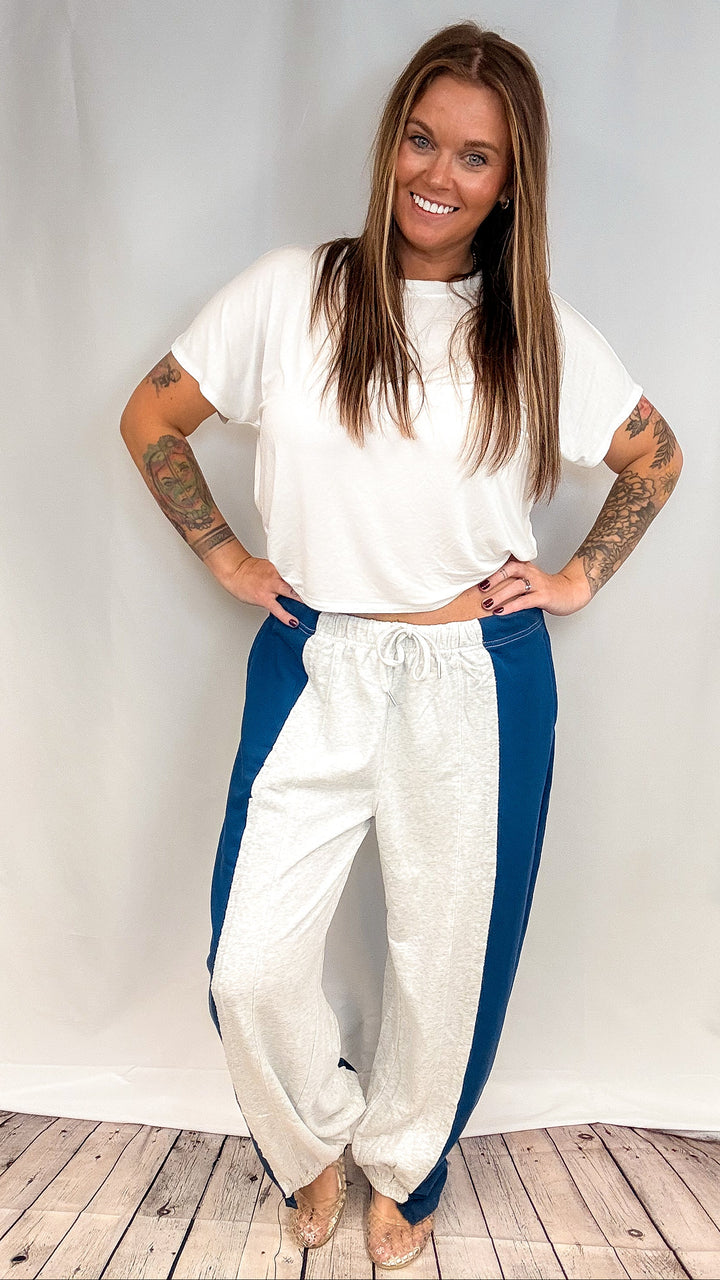 A woman modeling a white t-shirt and heathered white sweatpants with blue side panels, standing against a white fabric backdrop on a wooden floor.