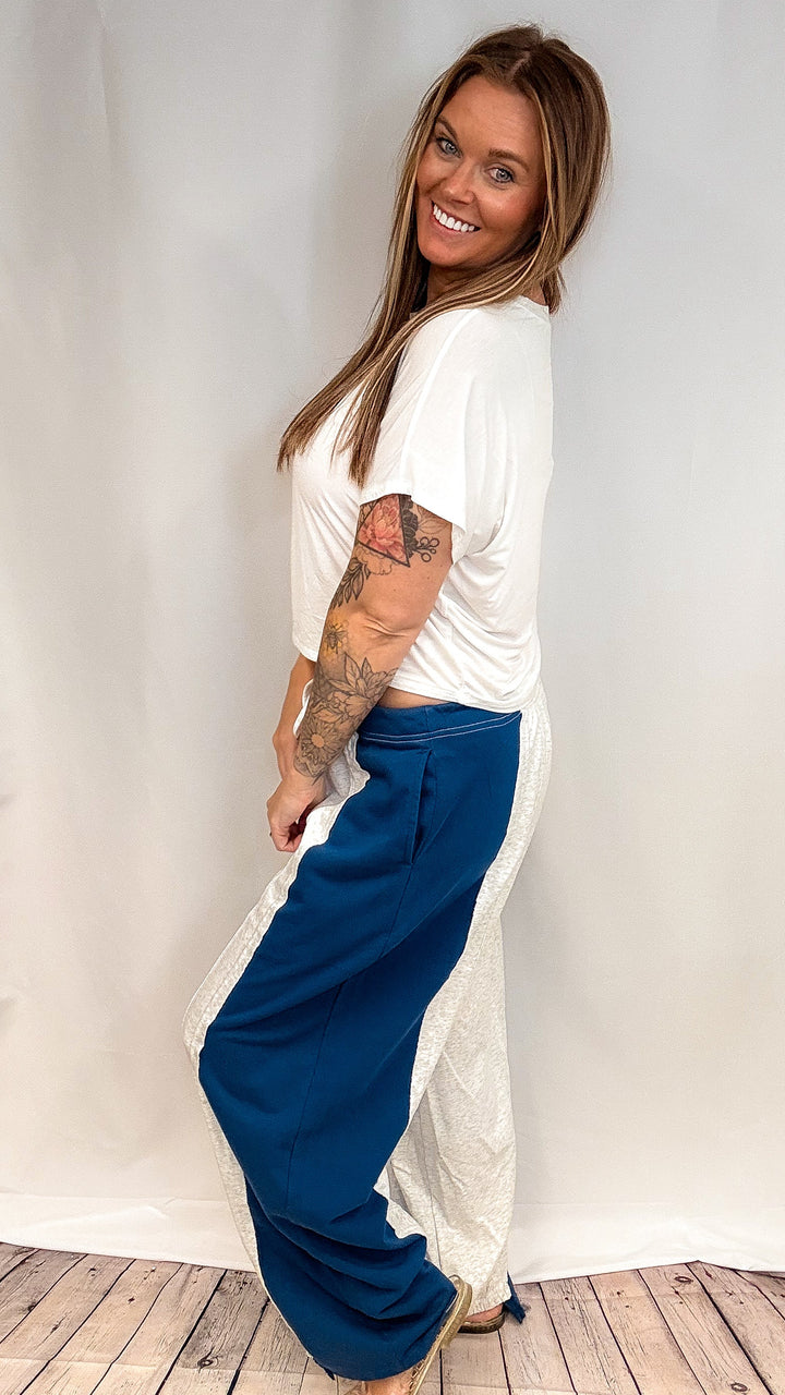 Woman wearing grey and navy sweatpants with short sleeve white top on plain background