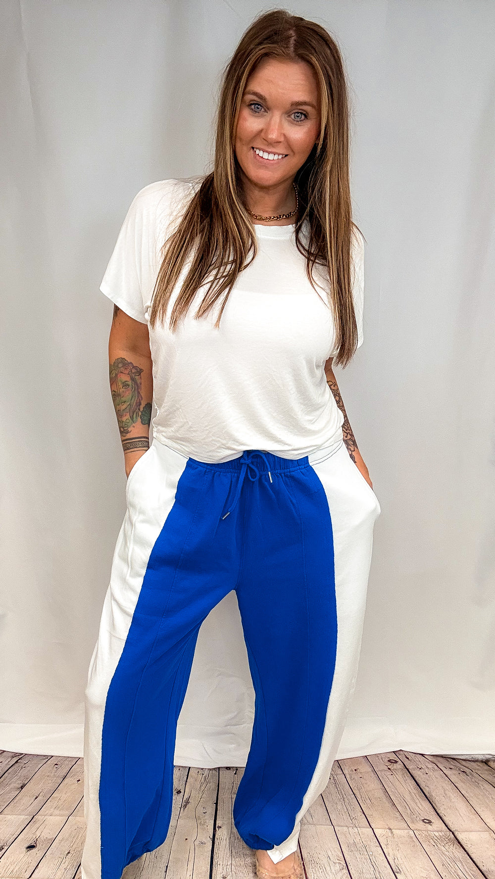 A woman modeling a white t-shirt and white wide-leg pants with vibrant blue side panels, standing against a white studio backdrop with a wood floor visible at the bottom.