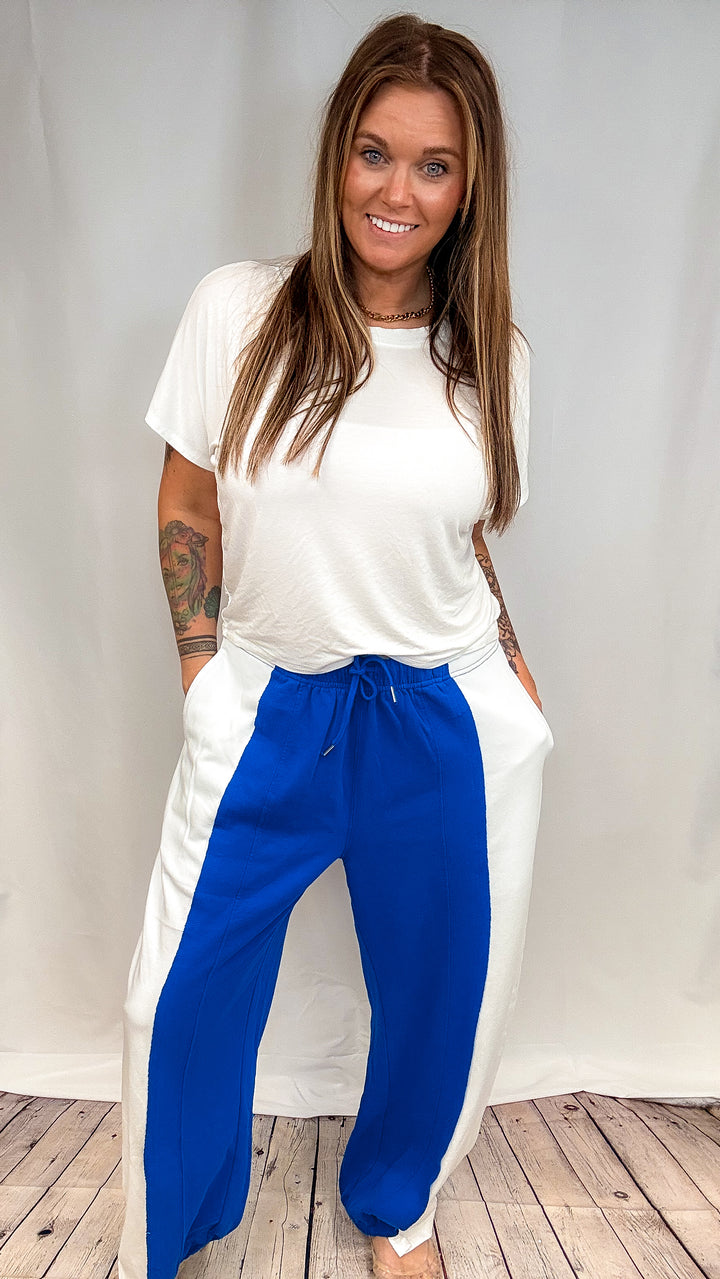 A woman modeling a white t-shirt and white wide-leg pants with vibrant blue side panels, standing against a white studio backdrop with a wood floor visible at the bottom.