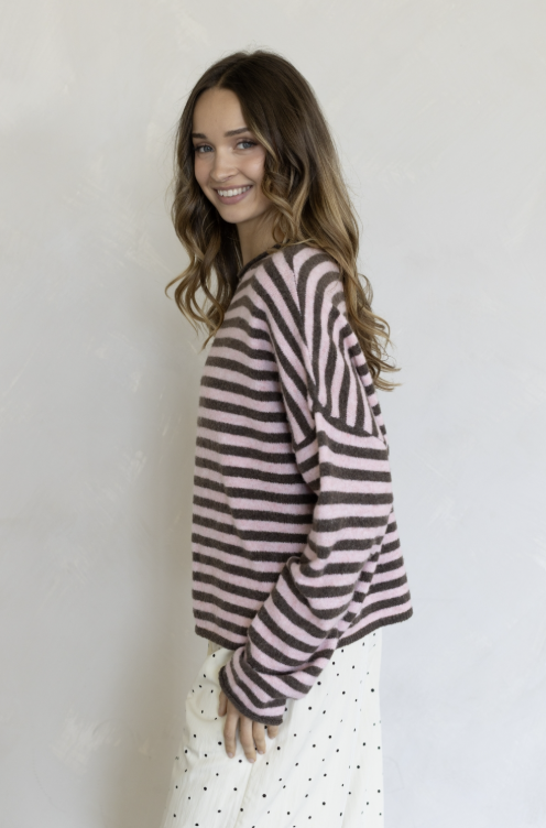Woman wearing pink and brown stripe pullover and white pants on plain background