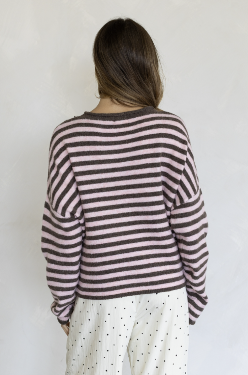 Woman shown from behind wearing pink and brown stripe pullover and white pants on plain background