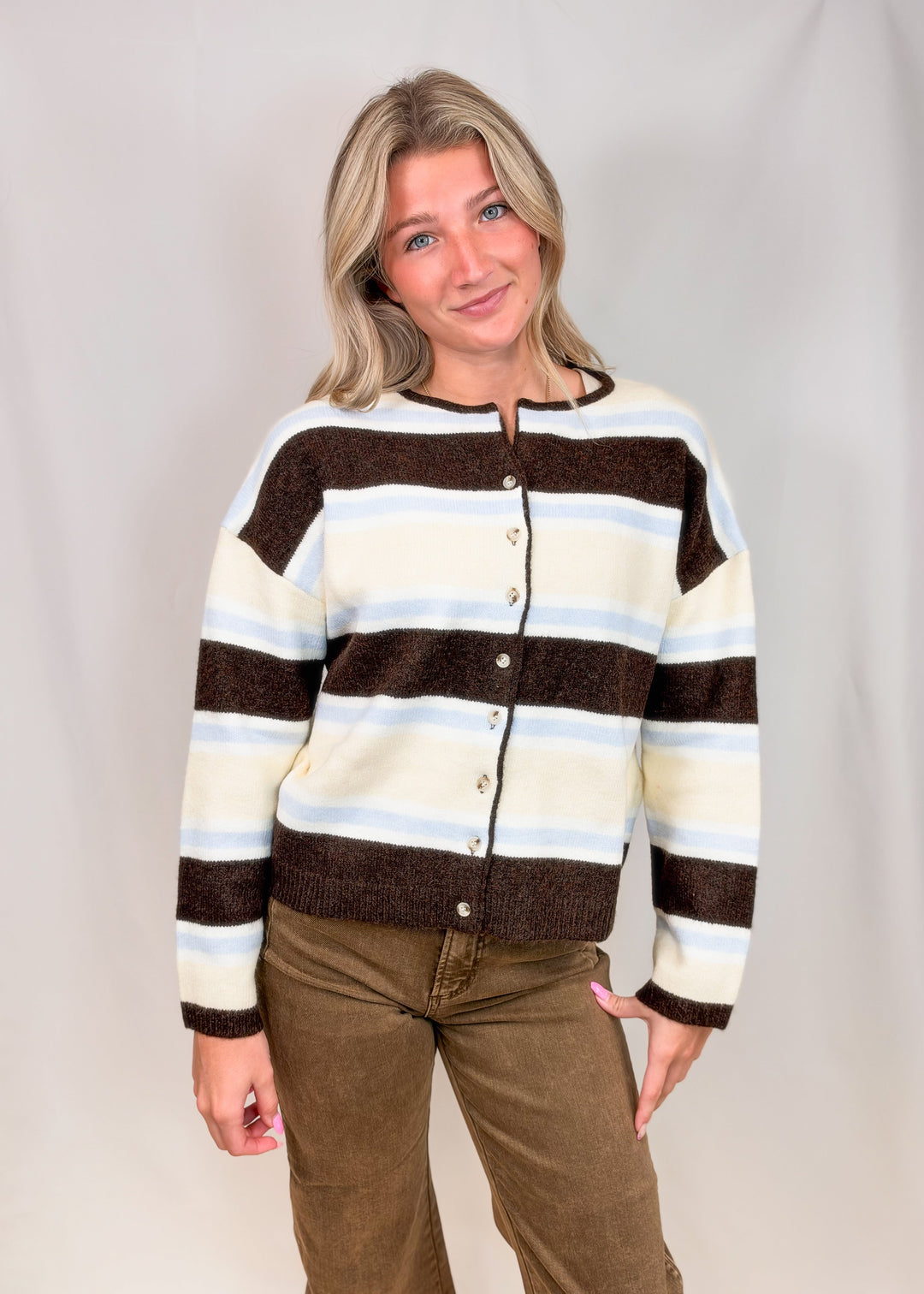 Woman wearing cardigan with brown, light blue and yellow stripes with brown pants on plain background