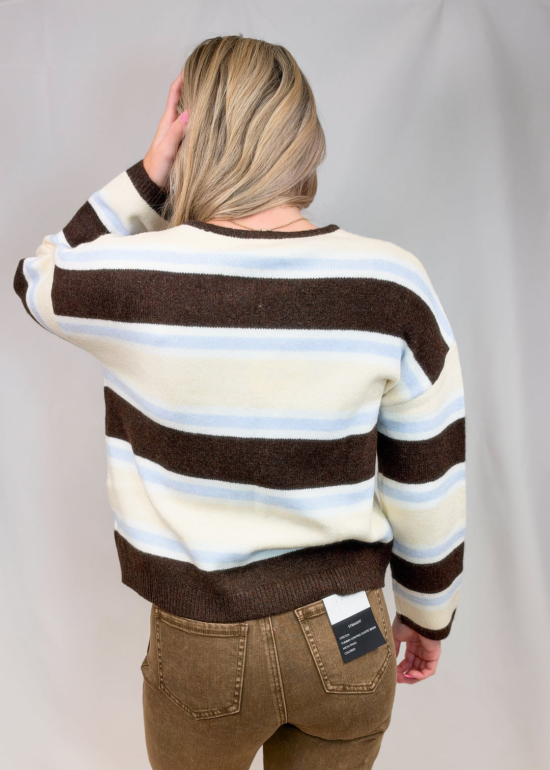 Woman shown from back view wearing brown, light blue and yellow stripe cardigan and brown pants on plain background