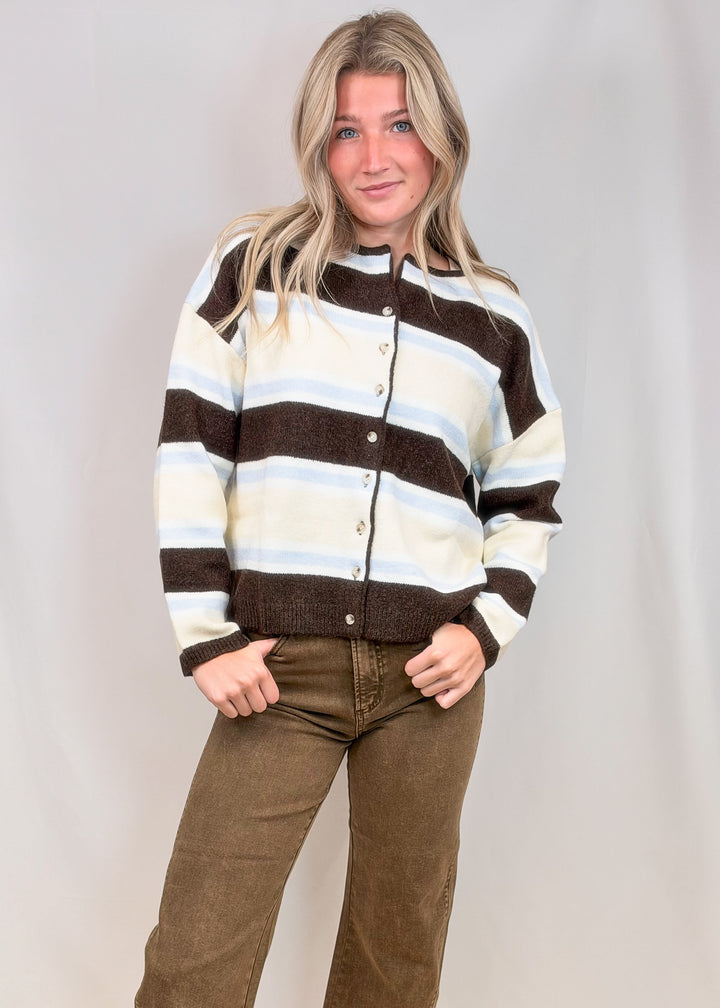 Woman wearing brown, light blue and yellow stripe cardigan with brown pants on plain background