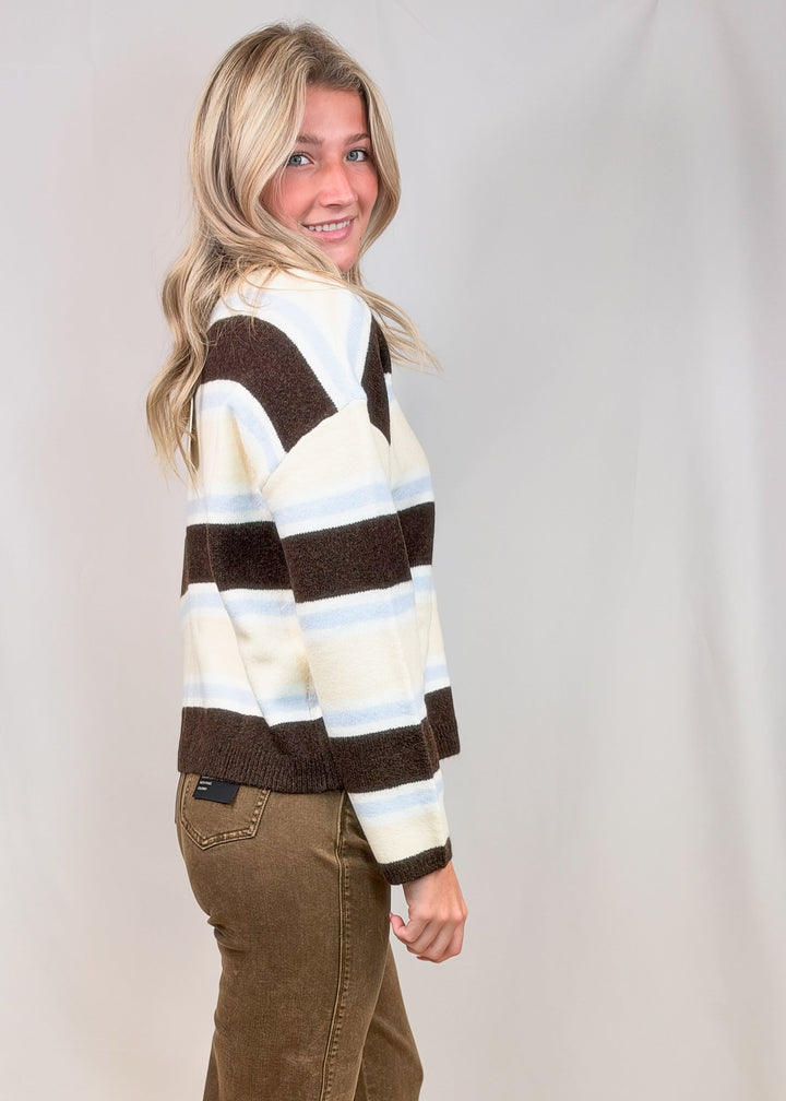 Woman shown from side view wearing brown, light blue and yellow cardigan with brown pants on plain background