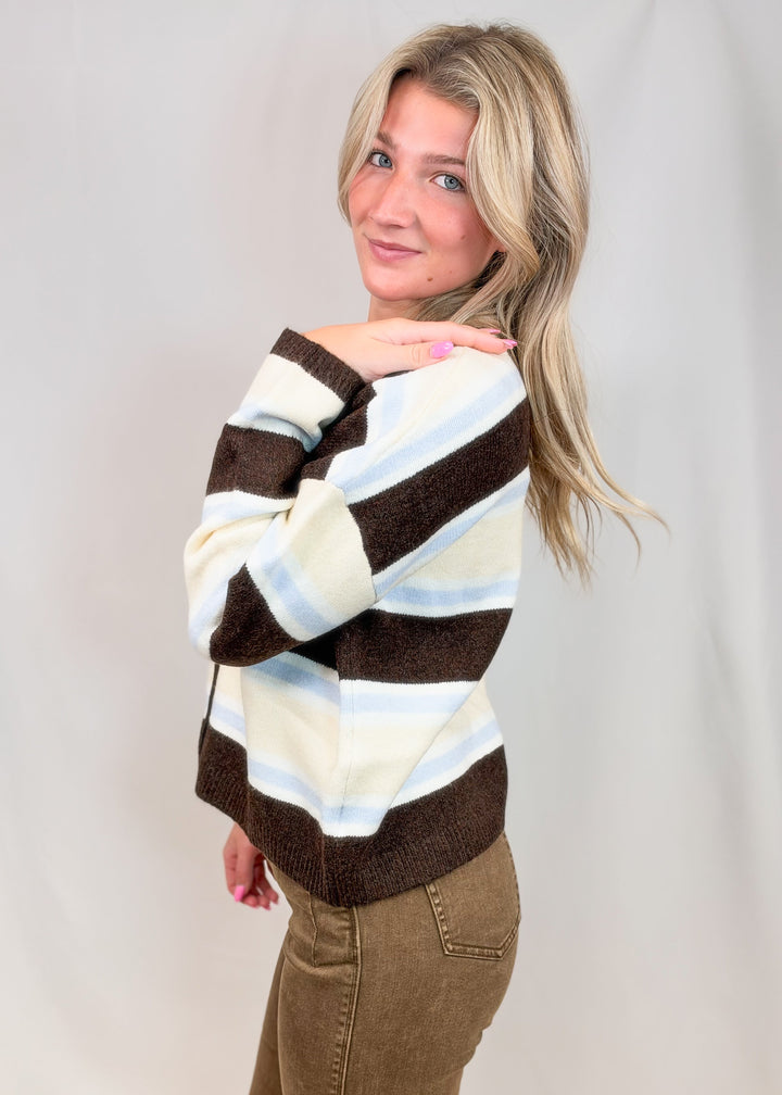 Woman shown from side view wearing brown, light blue and yellow stripe cardigan with brown pants on plain background
