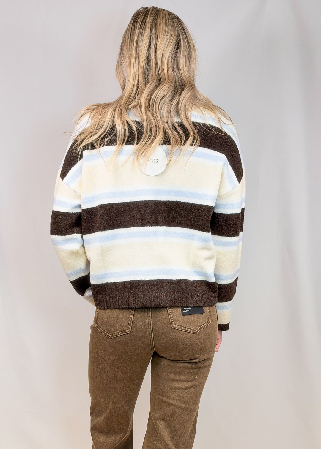 Woman shown from back view wearing brown, light blue and yellow stripe cardigan and brown pants on plain background