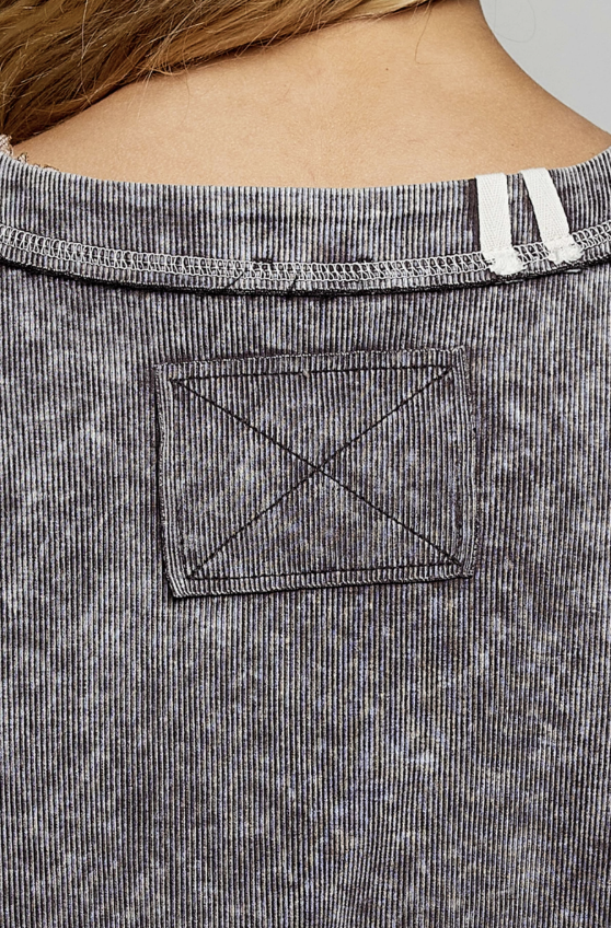 Close-up of the back of a ribbed gray top worn by a model, showing a square patch pocket with diagonal cross-stitching and white shoulder straps at the neckline.