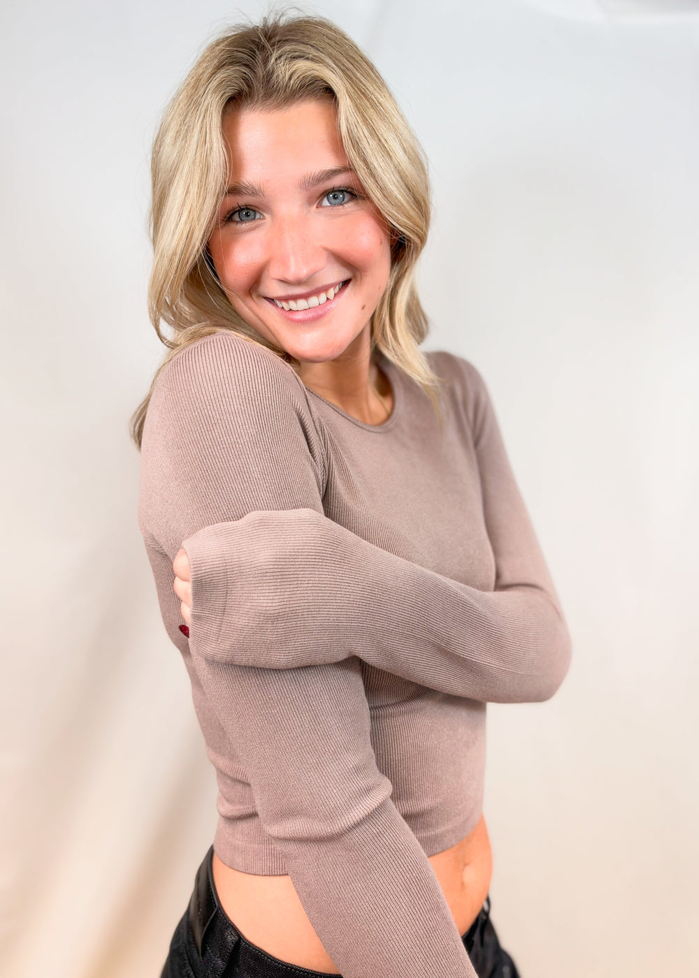 Woman wearing tan long sleeve top with black pants on plain background