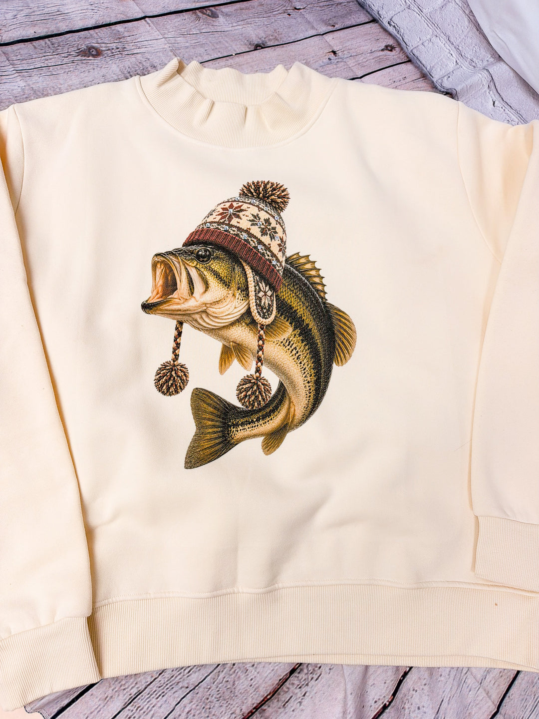 Cream-colored crewneck sweatshirt featuring a graphic of a largemouth bass wearing a knit winter hat with pinecone tassels, laid flat on a rustic wooden surface.