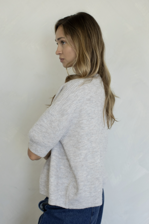 A woman with long light brown hair is shown from a side-back angle wearing a light gray heathered knit cardigan and blue jeans against a plain off-white wall.